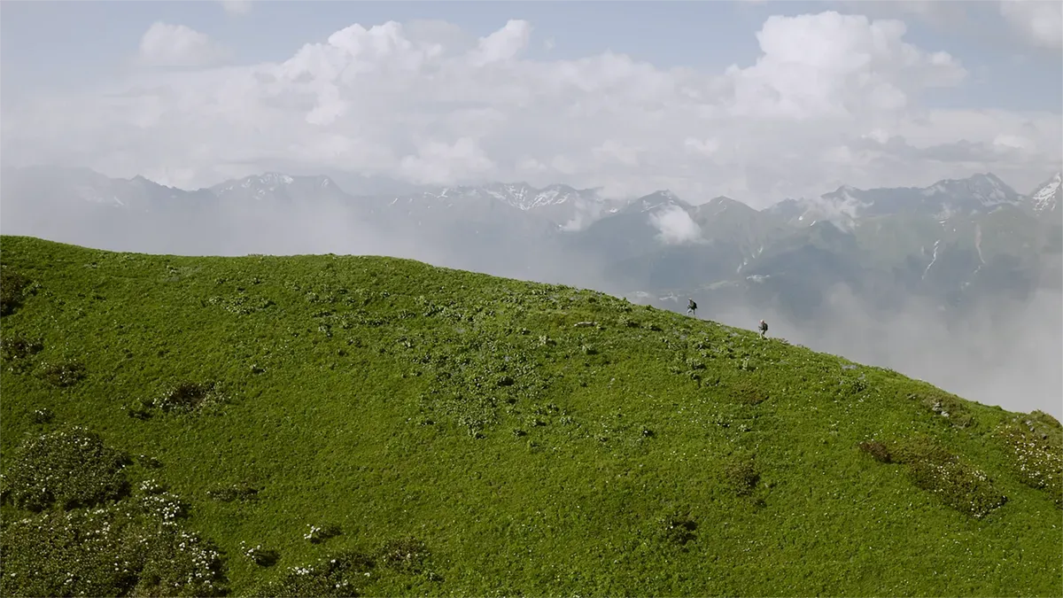 Two hikers walking on a lush green hillside with misty mountains and clouds in the background.