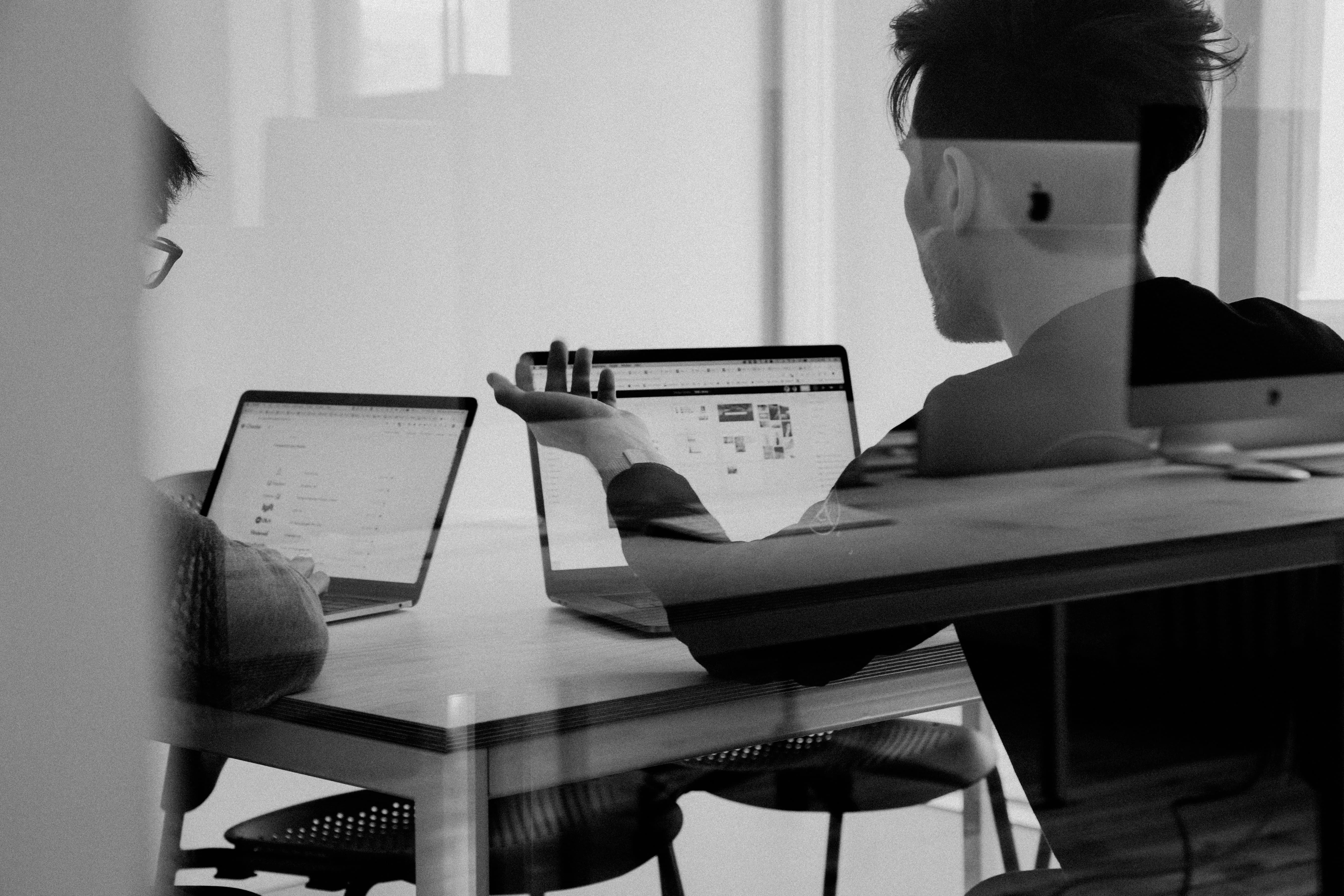 A black and white photo of two men sitting, working and talking at their laptops.