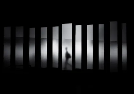 Black and white image of the figure of a man walking through a passage past a rectangular fragmented wall.